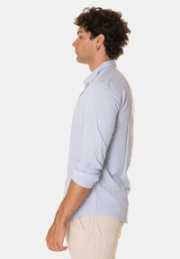 Camicia azzurra chiara con bottoni, maniche lunghe, dal taglio morbido e tessuto leggermente strutturato, vista di lato.