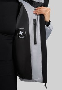 Interno di una giacca nera e grigia con tasca con zip, logo e texture liscia. Il design combina materiali e colori contrastanti.