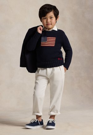Ungt barn iført en navyblå sweater med et amerikansk flag, hvide bukser, navy sneakers, holder en mørk jakke over den ene skulder.
