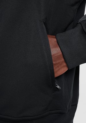 Hand mit dunkler Haut, die in eine schwarze, mit Reißverschluss versehene Tasche eines schwarzen Langarmshirts mit geripptem Bündchen gesteckt wird.