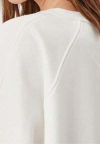 Tessuto di felpa bianca che mostra un primo piano della cucitura della spalla, con una consistenza morbida e un design a raglan. I dettagli delle cuciture sono appena visibili.