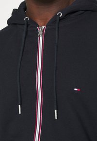 Sudadera con cremallera de color azul marino con rayas en rojo, blanco y azul a lo largo de la cremallera; cuenta con un logo en el pecho y acabados en metal plateado.