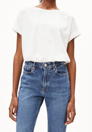 Personne portant une chemise blanche unie à manches courtes rentrée dans un jean bleu en denim taille haute avec poches avant et fermeture à boutons.