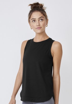 Woman with brown hair in a messy bun wearing gold hoop earrings and a sleeveless black top standing against a plain light background.