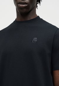 Zbliżenie osoby noszącej czarną koszulkę z okrągłym dekoltem z małym ciemnym haftowanym logo na lewej piersi.