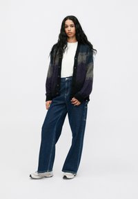 Moteris su ilgais garbanotais plaukais, vilkinti daugiaspalvį kardiganą, baltą marškinį, plačius džinsus ir baltus sportbačius, stovi prieš vienodą foną.