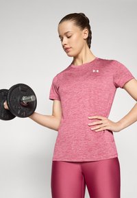 Camiseta deportiva de color rosa jaspeado con mangas cortas, combinada con leggins rosas brillantes. Sosteniendo una mancuerna negra con placas circulares.