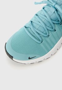 Lysblå træningssko med en struktureret stofoverdel, flade snørebånd og en hvid sål. Har et sort Nike-logo nær tåen.