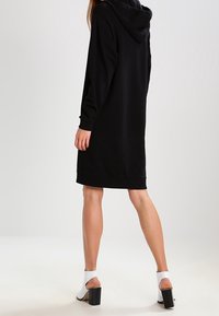 Svart huvtröja klänning med långa ärmar, rak passform och fåll runt mitten av låret. Bärs med vita klackskor med blockhæl.