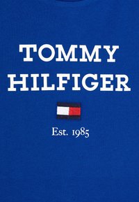 Modré bavlněné tričko s nápisem "TOMMY HILFIGER" v bílém písmu a vyšitým logo vlajky s červenými, bílými a modrými akcenty. Pod ním je uvedeno "Est. 1985".
