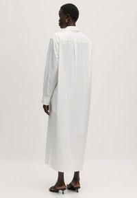 Weißes, langärmeliges Hemdkleid mit Kragen, gerader Silhouette und seitlichen Schlitzen. Glattes Material, bis zur Mitte der Wade reichend.