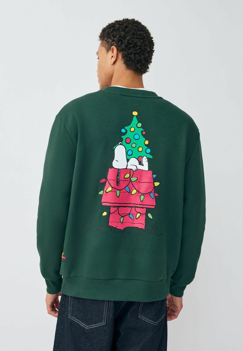 Zöld kapucnis pulóver, amelyen Snoopy látható egy rózsaszín kutyaházon, mellette egy díszített karácsonyfa és színes fények a háta mögött. Bordázott mandzsetta és alsó szegély.