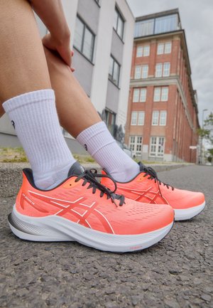 Person trägt leuchtend orange Laufschuhe und weiße Socken und sitzt auf dem Bordstein einer städtischen Straße mit Gebäuden im Hintergrund.