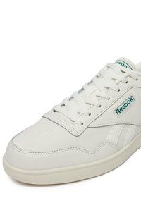 Białe skórzane trampki z okrągłym noskiem, teksturowaną powierzchnią, płaskimi sznurowadłami i zielonym akcentem logo. Posiadają przeszycia po bokach oraz gumową podeszwę.