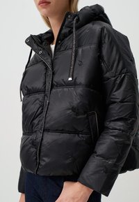 Zwarte pufferjack met een glanzende, gequiltte afwerking, voorzien van een capuchon, zilveren accenten en geborduurde details op de stof.