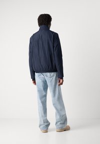 Veste de bombardier navy avec un col monté, des poignets élastiques et une taille élastique. Portée sur un jean bleu clair ample et des chaussures beige.