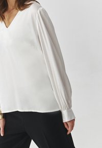 White silk blouse with a V-neck, long puff sleeves, and buttoned cuffs. Smooth texture with a slight sheen. Paired with black bottoms.