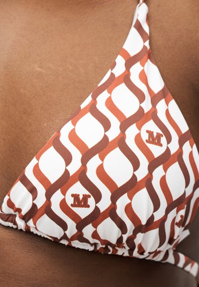 Κοντινή λήψη ενός τοπ μπικίνι με καφέ και λευκό μοτίβο, κυματιστές ρίγες και κεντητό γράμμα "M" πάνω σε σκούρο δέρμα.