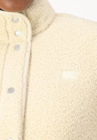 Kermanvärinen fleece-takki, jossa on korkea kauluspusero, hopeiset napit ja pieni logo-tagi etupuolella. Pehmeä rakenne ja mukava ulkonäkö.