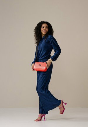 Navyblauwe geplooide co-ordset bestaande uit een lange blouse en bijpassende broeken. Geaccentueerd met een felrode rechthoekige handtas en roze hakken.