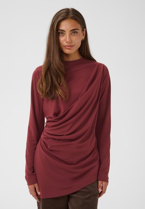 Femme aux longs cheveux bruns portant un haut bordeaux à manches longues avec un détail de tissu drapé et un pantalon marron, debout devant un fond uni.