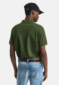 Man in een donker groene polo met korte mouwen, lichtblauwe jeans met een bruine riem en een donkerblauwe baseballcap, gezien vanuit de rug.