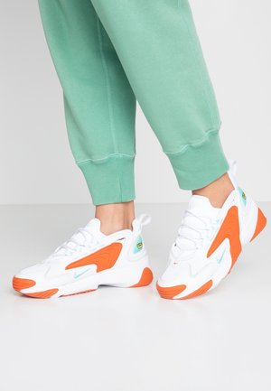 Hvide sneakers med orange accenter båret med grønne opsmøgede bukser, vist fra siden og foran på en ensfarvet hvid baggrund.