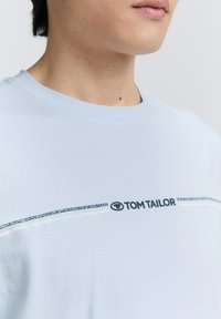 Šviesiai mėlynas marškinėliai iš minkštos medžiagos su apykakle ir horizontaliu grafiniu juosteliu, kuriame yra tamsios spalvos tekstas "TOM TAILOR".