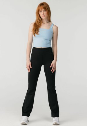 Punatukkainen nainen, jolla on vaaleansininen hihaton crop-top, mustat istuvat housut, valkoiset lenkkarit ja kaulakoru, seisoo yksinkertaisen taustan edessä.