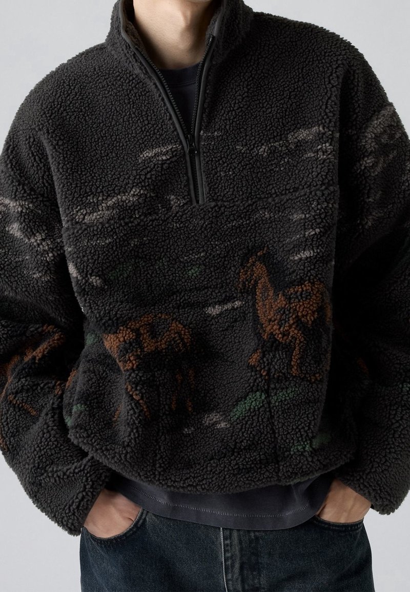 Personne portant un pull polaire gris foncé avec une fermeture éclair quart de tour et un motif abstrait marron et vert sur le devant et les manches.