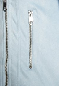 Primo piano di una giacca di tessuto blu con una cerniera argentata dotata di un caratteristico tirante, e dettagli di superficie testurizzati. Aspetto liscio e elegante.