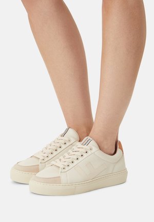 Pieds portant des baskets basses crème avec des accents beige et tan, lacées, sur un fond blanc uni.
