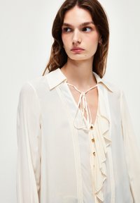 Blusa de crema en tejido ligero con sutiles detalles de volantes, acentos de botones, escote con lazo y mangas largas. Finas pletas verticales en la parte delantera.