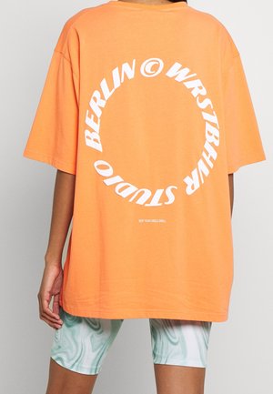 Osoba nosząca za dużą pomarańczową koszulkę z białym okrągłym napisem "Berlin Wrstbhr Studio" i zielone marmurkowe szorty, stojąca na jednolitym tle.