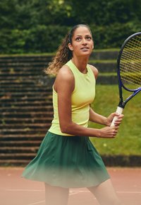 Draag een neongele mouwloze top en een groen-gradiënt tennisrok, met een racket in hand op een tennisbaan met gras op de achtergrond.