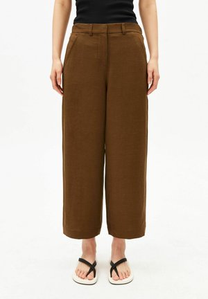 Personne portant un pantalon large marron court, un haut noir sans manches et des sandales blanches à brides noires, debout sur un fond blanc.