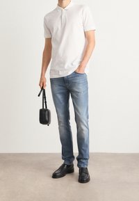 Camisa polo blanca de manga corta, vaqueros azules desteñidos y zapatos de cuero negros. Sosteniendo una pequeña bolsa negra con una correa.