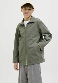 Olivgrön zip-up jacka med spetsig krage, två framfickor och en jämn textur, bärs över en vit skjorta och grå byxor.