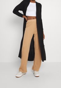 Schwarzer langer Cardigan, beige gerippte weit geschnittene Hose, weißes kurzes Oberteil und weiße Sneaker. Besitzt Textur und kontrastierende Farben.