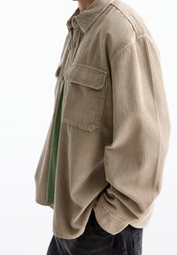 Camisa de pana beige con un corte holgado, dos bolsillos delanteros, cierre de botones y una superficie texturizada sobre una capa base verde.