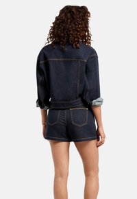 Veste en denim foncé avec les manches retroussées, ornée de coutures jaunes contrastantes, associée à un short en denim foncé assorti avec des poches arrière.