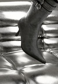 Juodos odos aukštakulniai batai su smailiu pirštu, dekoratyviniais sagtelėmis ir klijuotomis diržais, padėti virš blizgaus metalinio paviršiaus.