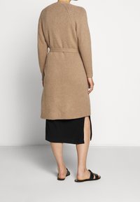 Beżowy kardigan dzianinowy z długimi rękawami i paskiem, z wykończeniem w prążki. Połączony z czarną spódnicą z bocznym rozcięciem i czarnymi sandałami.