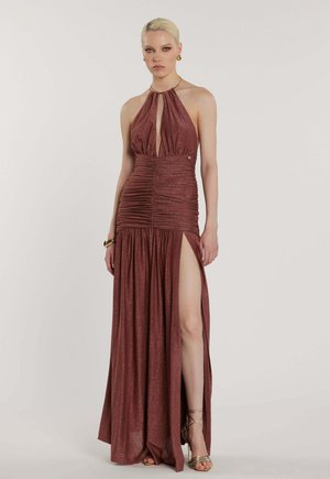 Donna che indossa un abito senza maniche, scintillante color ruggine con vita arricciata, scollatura profonda a V e spacco alto, accessoriato con gioielli dorati e tacchi.