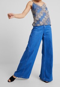 Camisola blu a fantasia con spalline sottili, abbinata a pantaloni in denim blu a gamba larga. Tessuto texturizzato e vestibilità ampia. Tacchi neri open-toe.