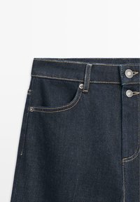 Ciemne dżinsy z denimu o średnim stanie. Posiadają zapinany na guziki, dwie przednie kieszenie oraz kontrastowe żółte przeszycia.