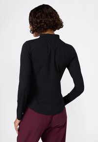 Maglietta nera a maniche lunghe con collo alto, texture a coste, orlo curvo e dettaglio di cucitura lungo la schiena, abbinata a pantaloni color marsala.
