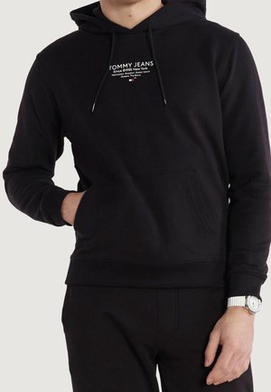 Person wearing black Tommy Jeans hoodie with front pocket and white watch on left wrist, hand in pocket, neutral background.