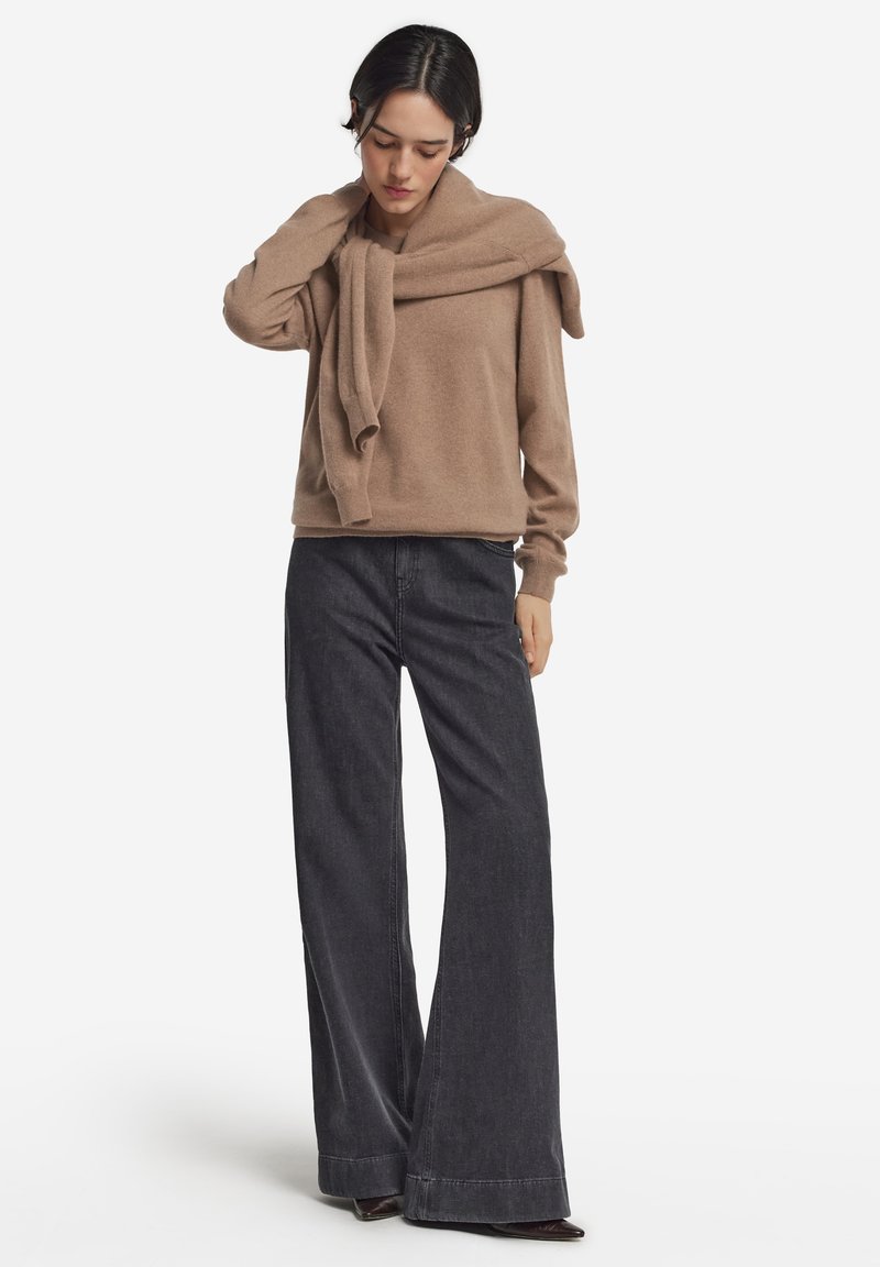 Hellbrauner Pullover mit weitem Ausschnitt und langen Ärmeln, über dunklen, weiten Jeans. Weiche Textur, minimalistisches Design und lässige Passform.