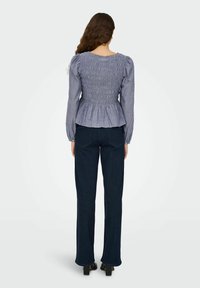 Blaue langärmlige Peplum-Bluse mit gerafftem Detail auf dem Rücken, kombiniert mit dunklen Jeans mit Schlag und Schuhen mit Blockabsatz.